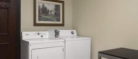 A laundry room with two stacked washers and dryers, a framed landscape painting on the wall, and a wooden door.