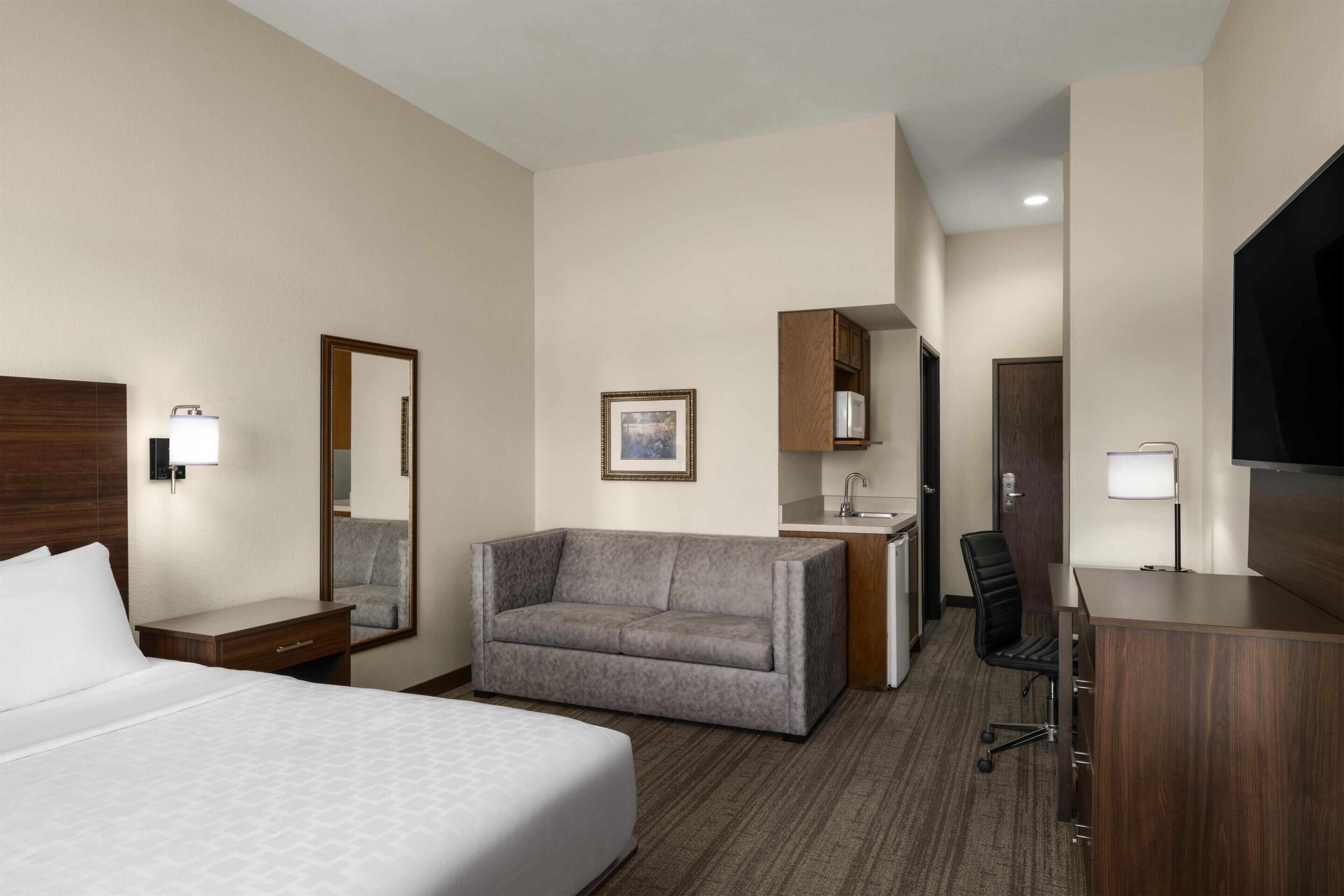 A hotel room with a bed, a sofa, a desk, a chair, a TV, a mirror, and a small kitchenette.