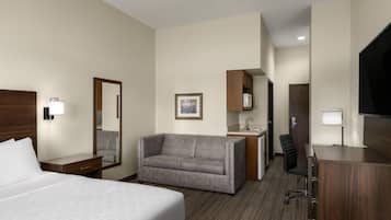 Habitación de hotel con cama, sofá, escritorio, silla, televisor, espejo y una pequeña kitchenette.