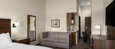 A hotel room with a bed, a sofa, a desk, a chair, a TV, a mirror, and a small kitchenette.