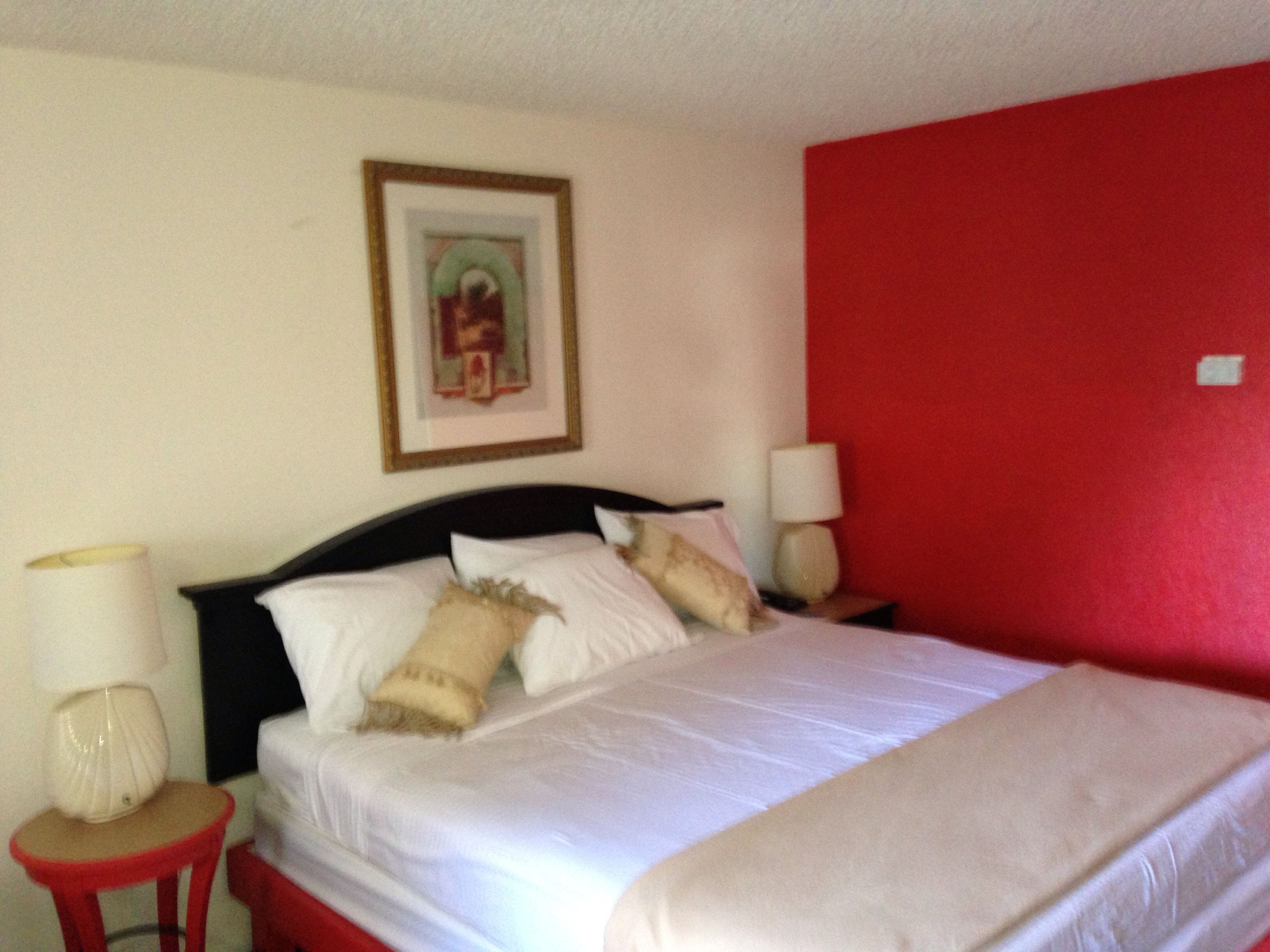 Una cama con sábanas blancas, una pared roja y un cuadro en la pared.