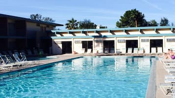 Una piscina con agua cristalina y azul, rodeada de sillones de descanso y un edificio con varias puertas y ventanas.