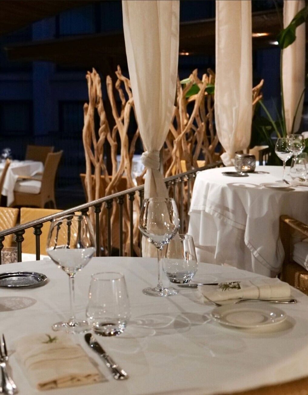Un tavolo da pranzo apparecchiato con tovaglie bianche, posateria e bicchieri da vino, circondato da elementi decorativi naturali e una scala.