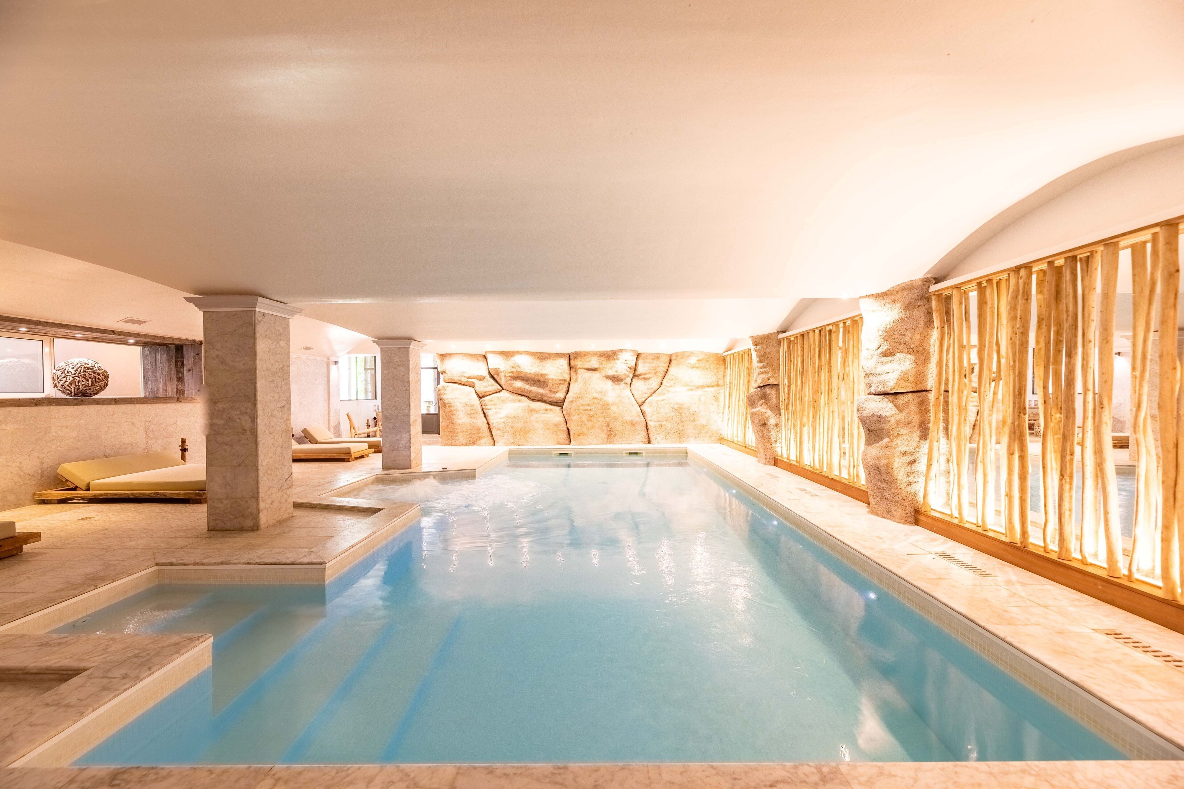 An indoor swimming pool with a stone wall backdrop, a seating area with cushions, and a wooden fence.