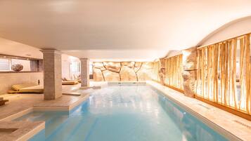 An indoor swimming pool with a stone wall backdrop, a seating area with cushions, and a wooden fence.