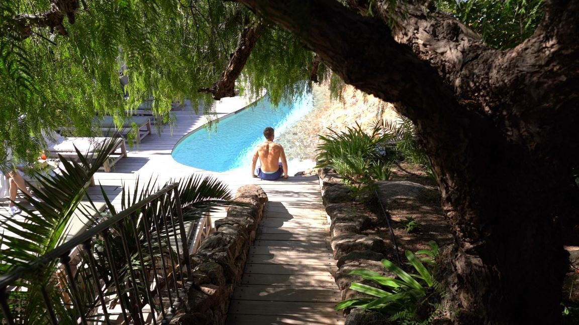 A man sitting by a pool with a wooden deck and stone walls, surrounded by greenery.