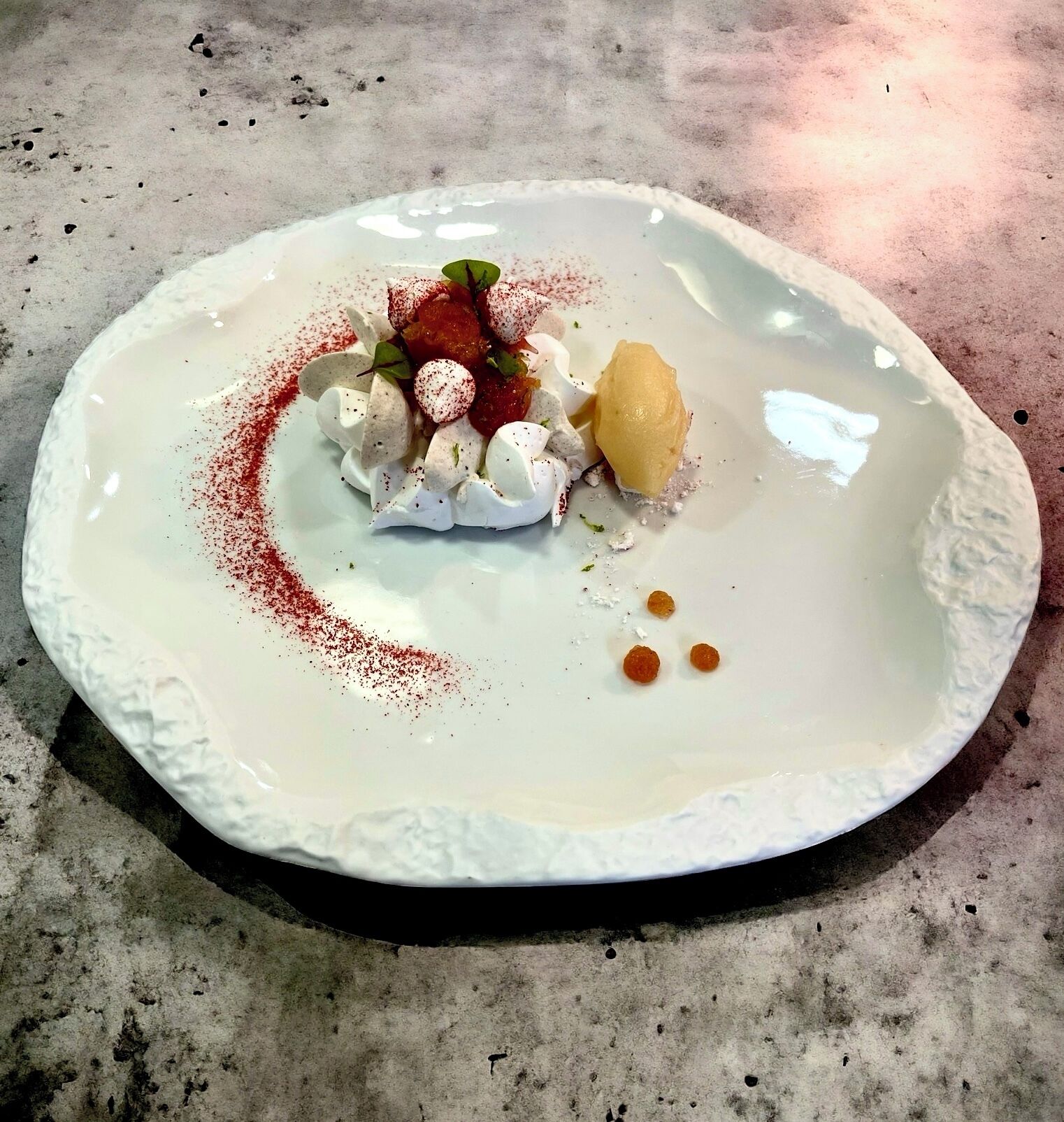 Ein Dessert auf einem weißen Teller mit strukturierter Kante, bestehend aus Sahne, Früchten und einer roten Puderzugabe.
