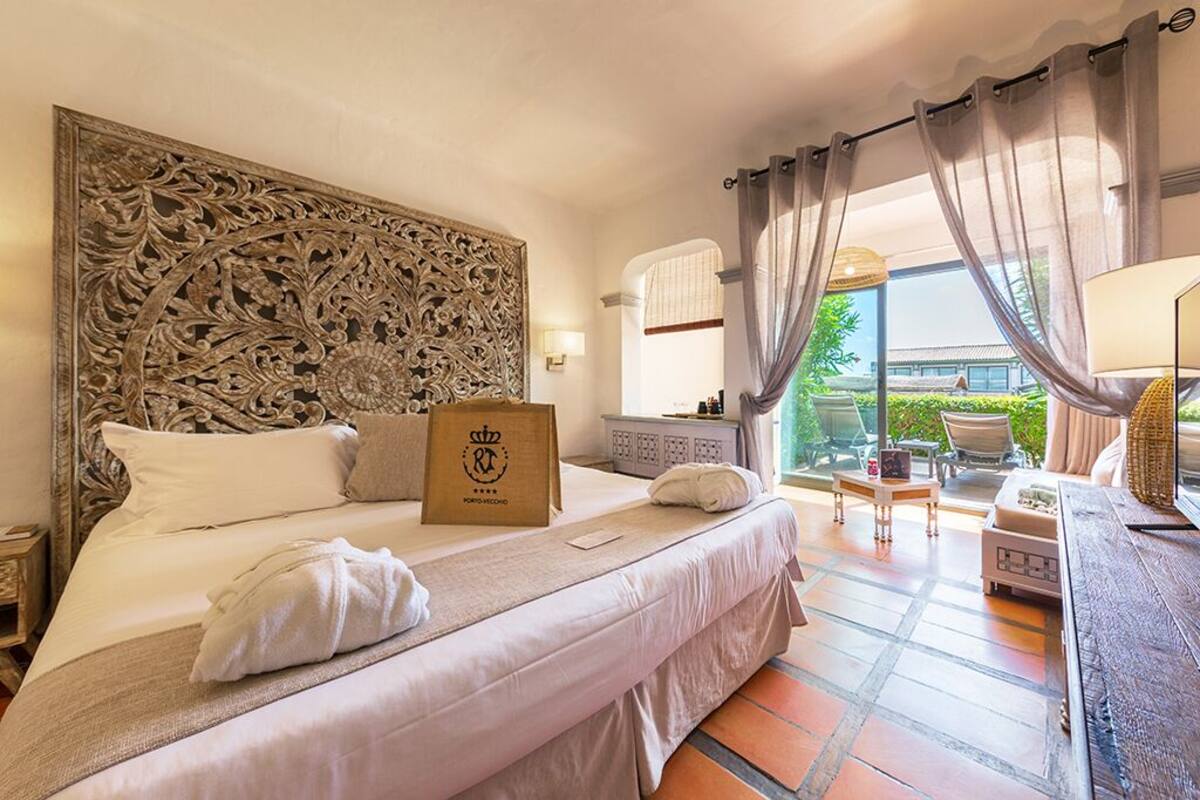 A hotel room with a large ornate headboard, a bed with white linens, a wooden nightstand, a suitcase, and a balcony with outdoor furniture.