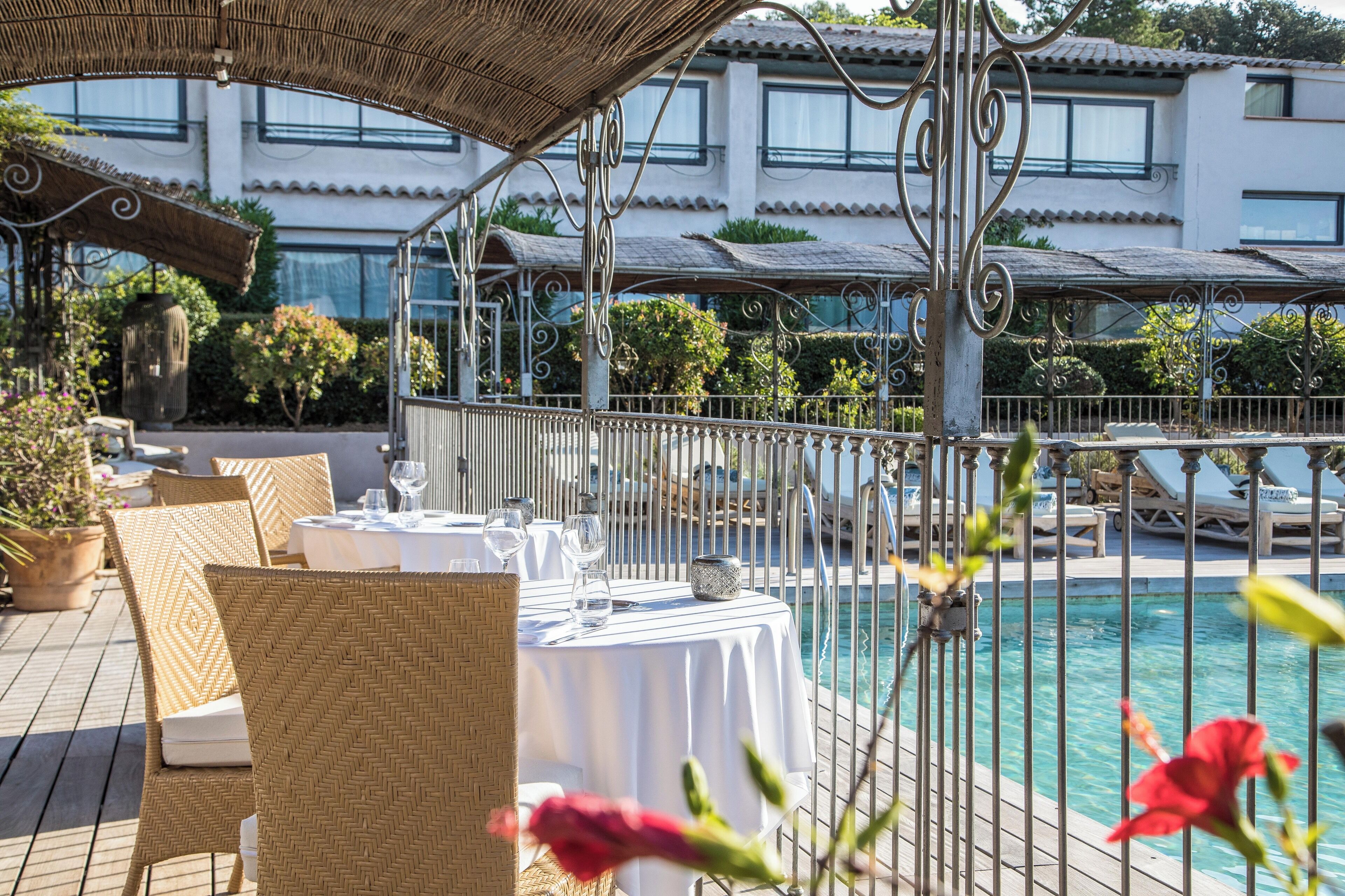 Un espace extérieur aménagé pour les repas, avec des chaises et des tables en osier, une piscine et, au fond, un bâtiment doté de balcons.