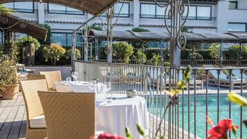 Un espace extérieur aménagé pour les repas, avec des chaises et des tables en osier, une piscine et, au fond, un bâtiment doté de balcons.