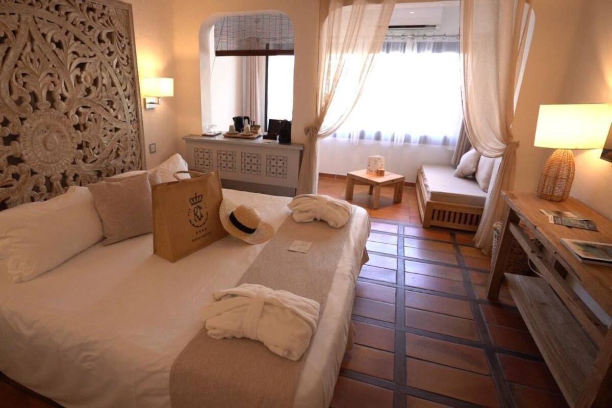 A hotel room with a large bed, a wooden headboard, a bedside table with a lamp, a small table with a chair, and a window with curtains.
