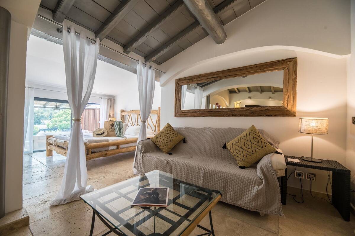 A cozy room with a wooden bed, a glass table with a book, a sofa with cushions, a mirror, and a lamp.