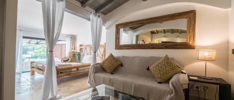 A cozy room with a wooden bed, a glass table with a book, a sofa with cushions, a mirror, and a lamp.