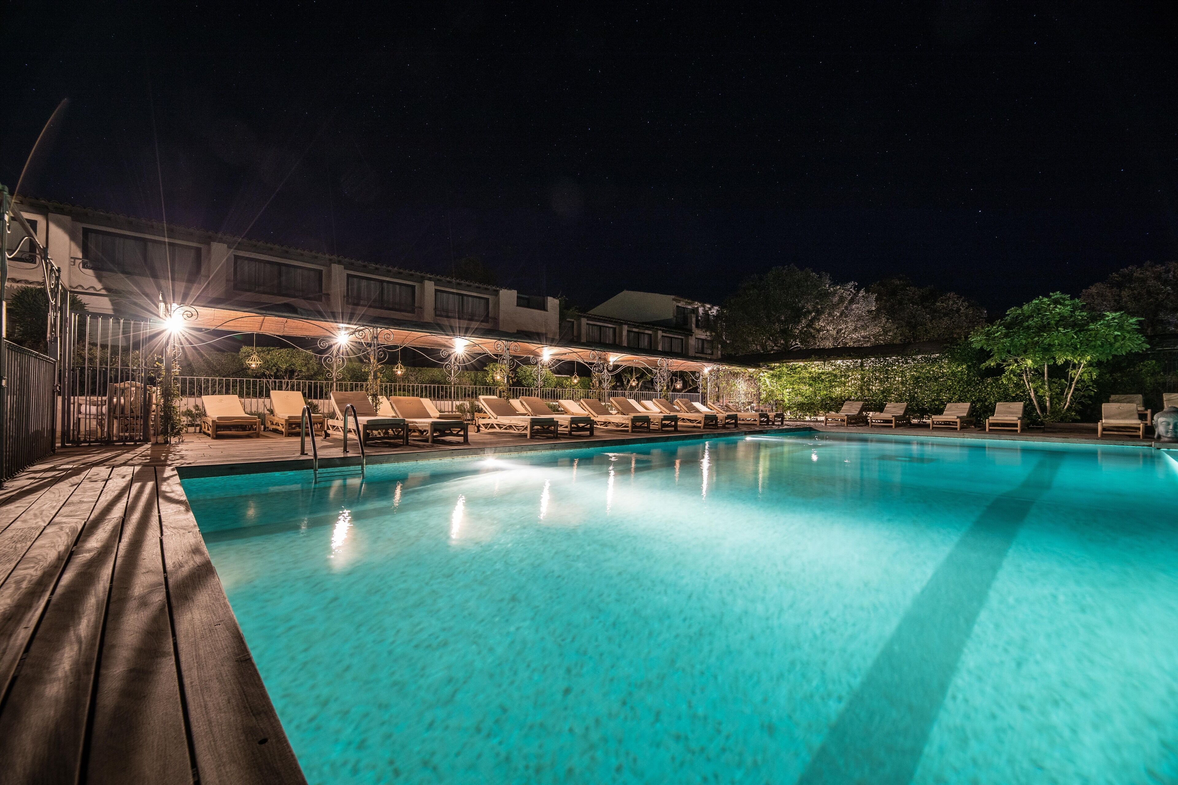 Ein Pool mit Liegestühlen und einer überdachten Liegefläche, umgeben von einer Holzterrasse und einem Gebäude mit zahlreichen Fenstern.