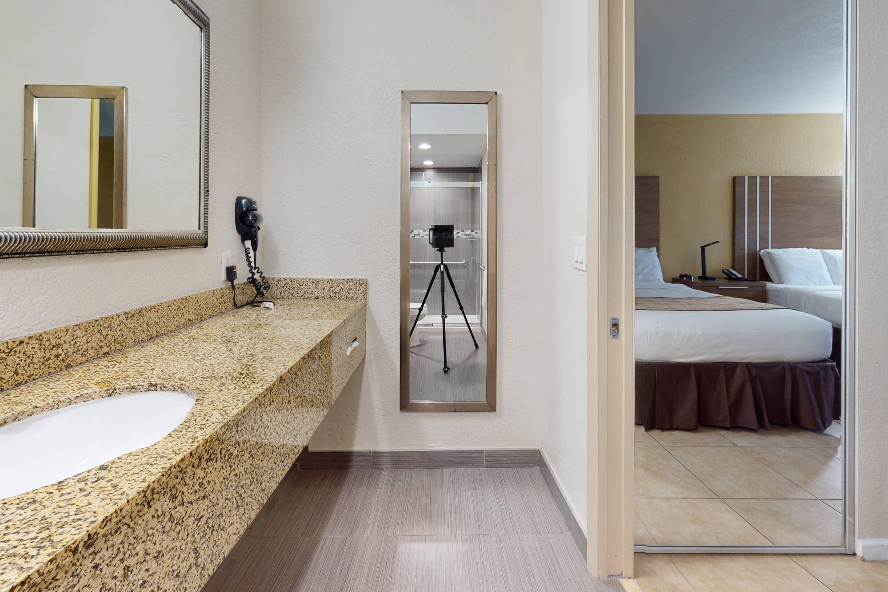 Un baño de hotel con encimera de granito, un lavamanos, un secador de pelo y una cámara con trípode.