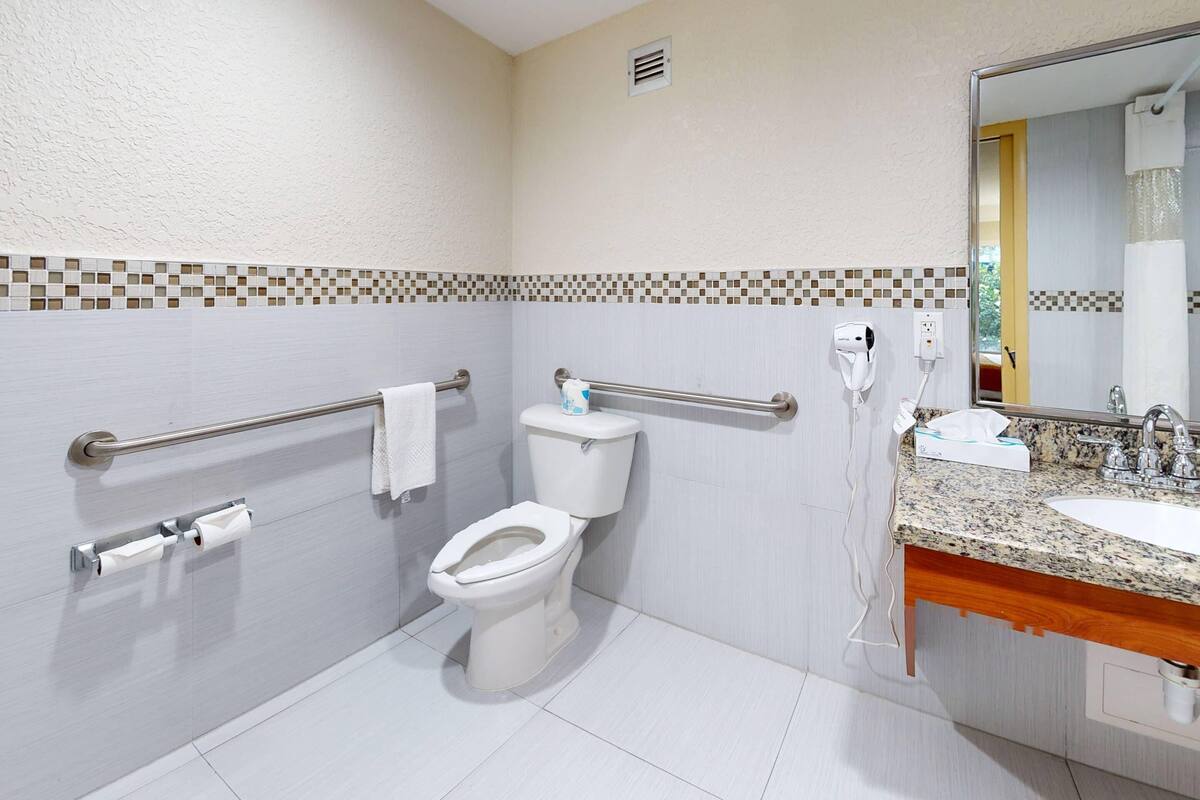 Een badkamer met een wit toilet, een handgreep, een handdoekhouder, een wastafel met spiegel en een betegelde spatwand.