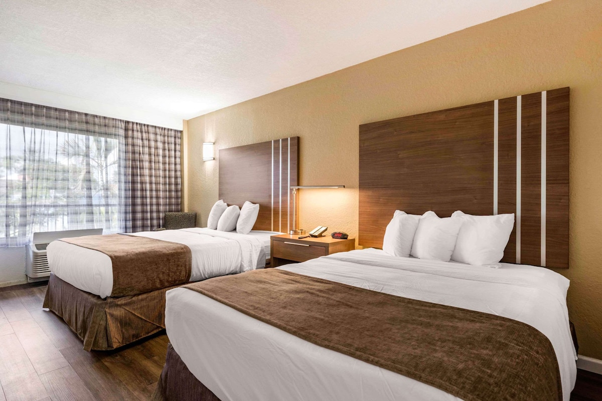 Een hotelkamer met twee bedden, een groot raam met gordijnen, een nachtkastje met een lamp en een aan de muur bevestigde televisie.