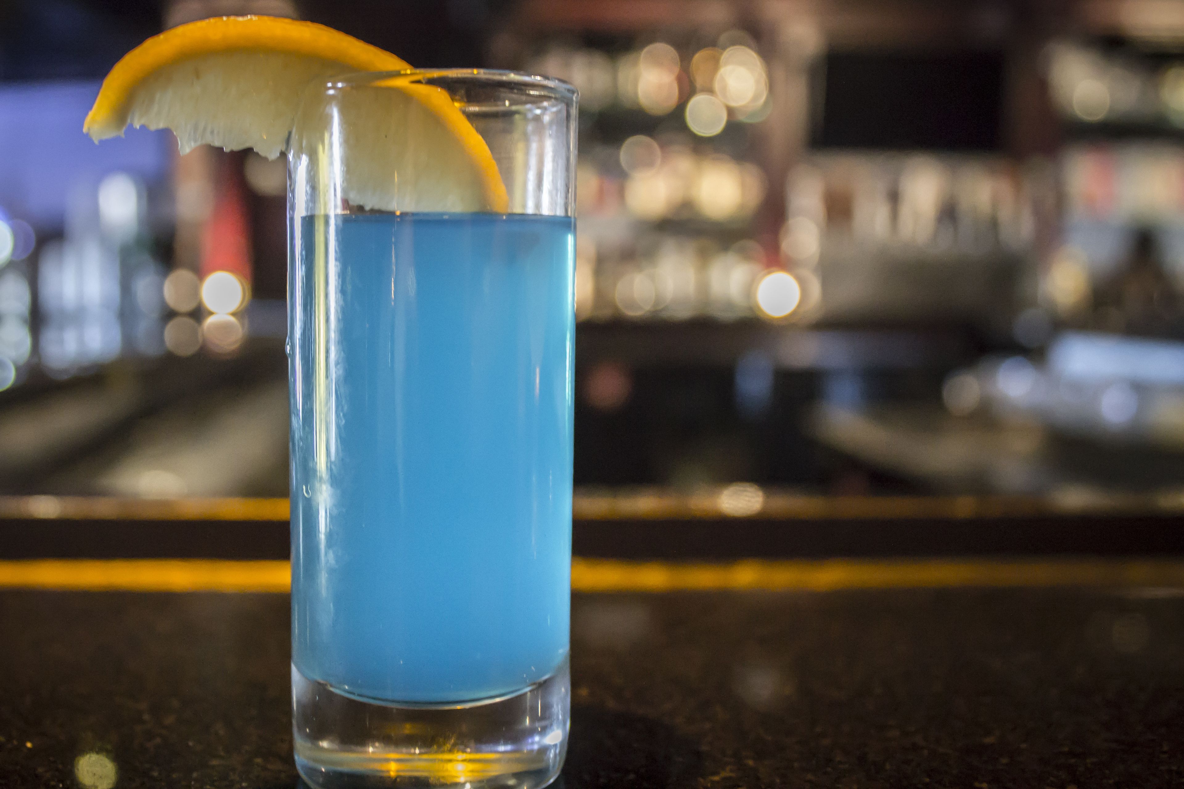 Un verre transparent contenant un liquide bleu, agrémenté d’une tranche d’orange et d’un élément blanc, qui pourrait être un citron, posé sur le comptoir d’un bar.