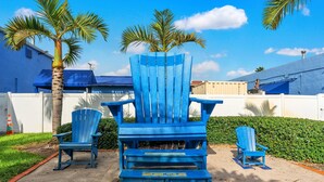 Een aantal blauwe Adirondack-stoelen in een zandrijk buitengebied, met een palmboom en een haag op de achtergrond.