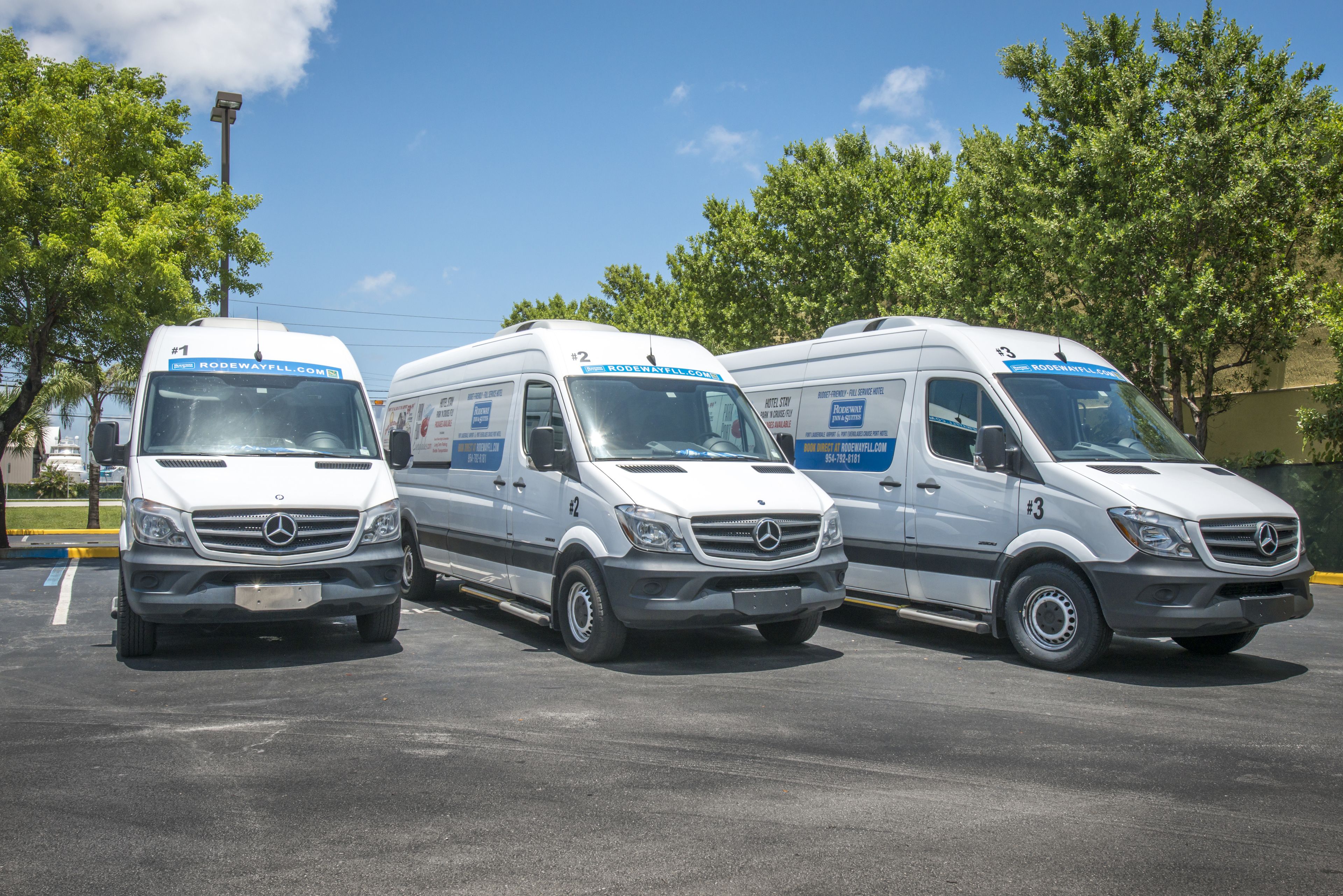 Three white Mercedes-Benz vans parked in a lot with trees and a clear sky in the background.