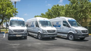 Trois fourgonnettes Mercedes-Benz blanches sont stationnées sur un parking bordé d’arbres, avec un ciel dégagé en arrière-plan.