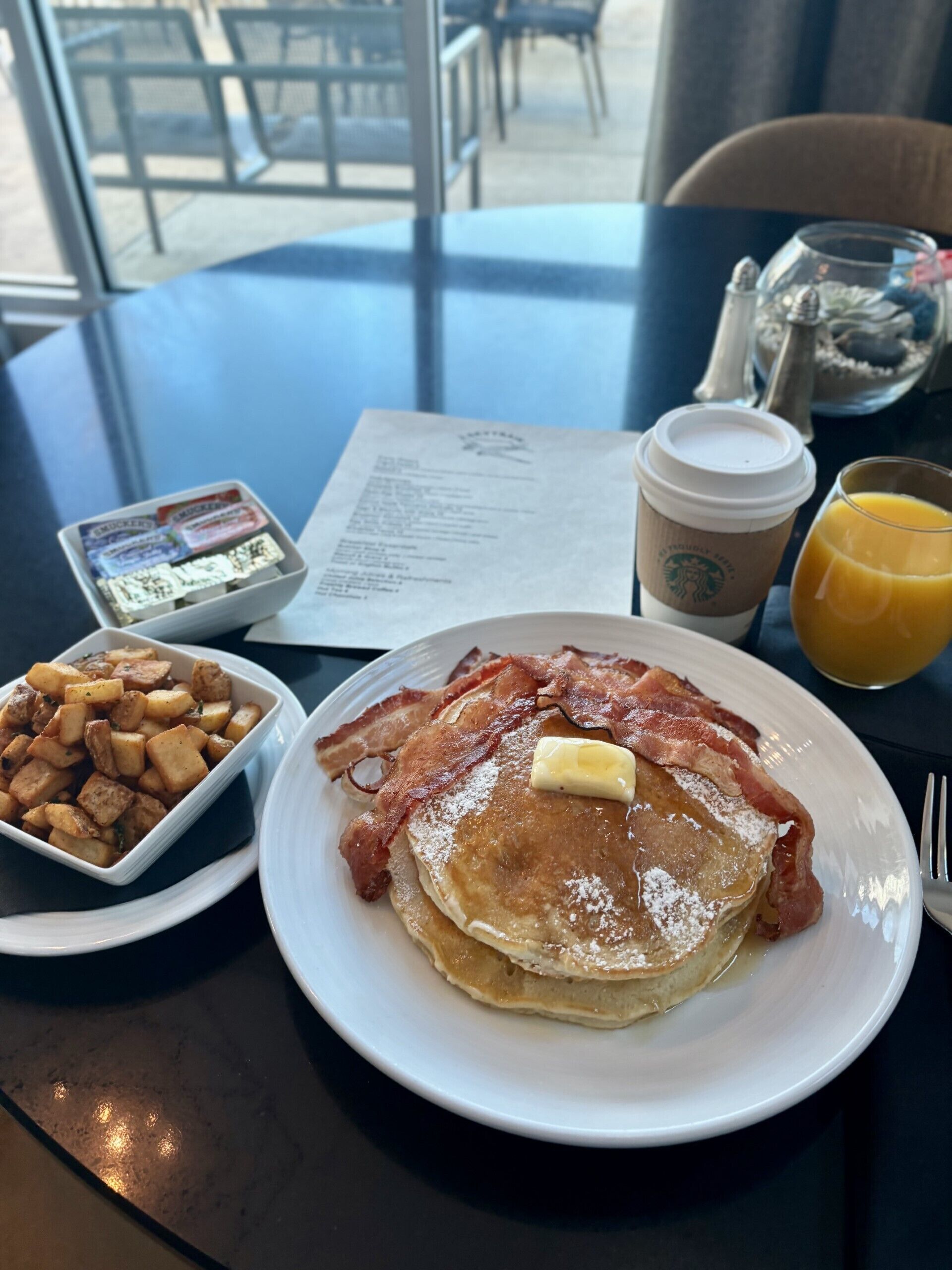 A breakfast table with pancakes, bacon, hash browns, a coffee cup, and a glass of orange juice.