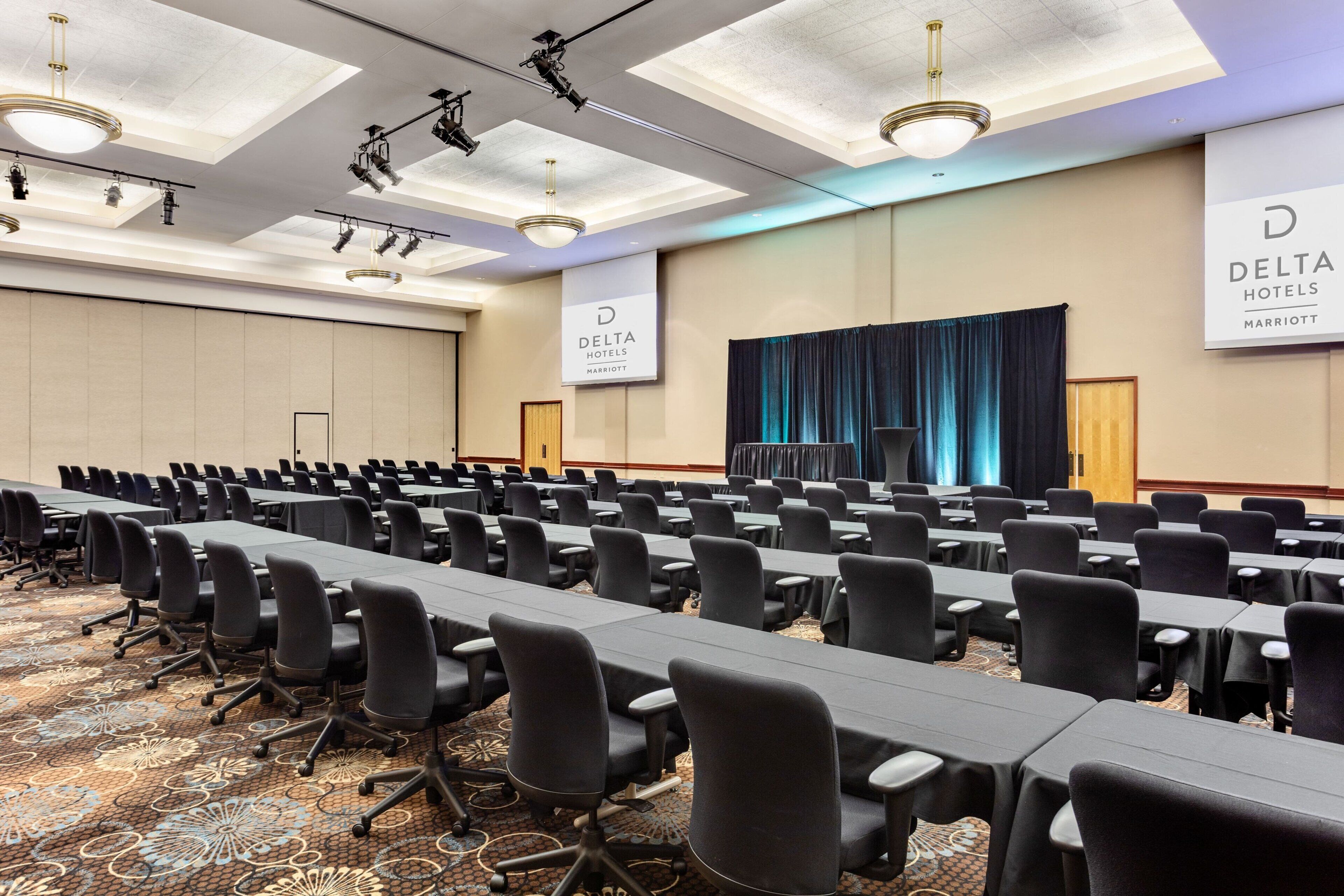 A conference room with rows of tables and chairs, ceiling lights, and banners for Delta Hotels Marriott.