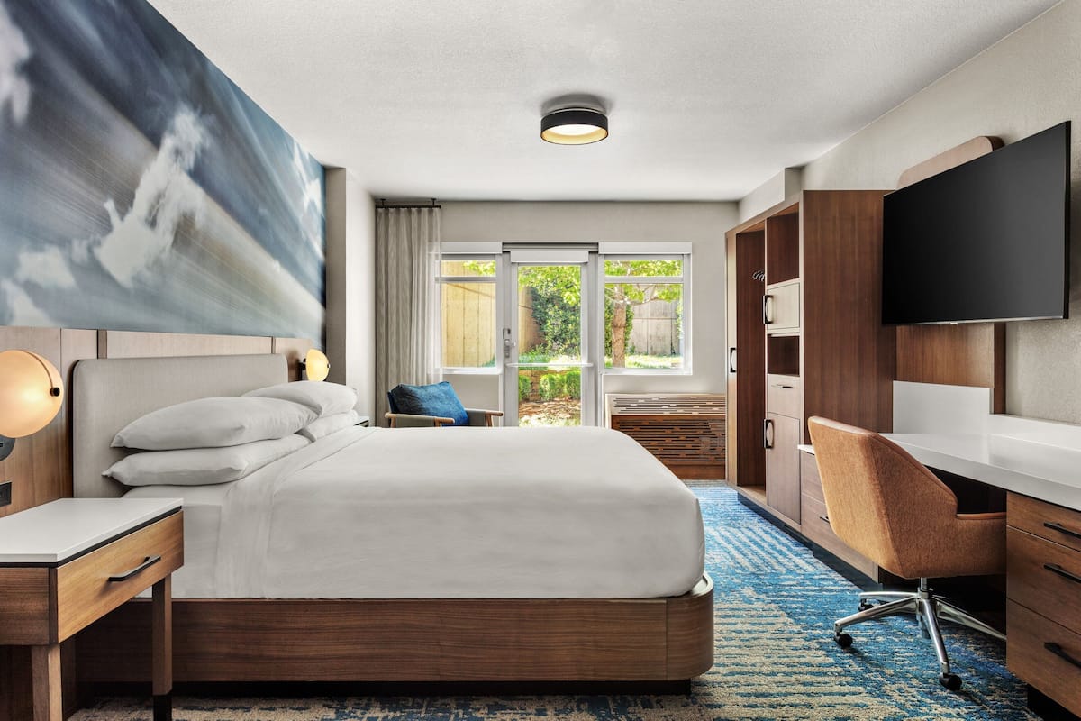 Habitación de hotel con cama, escritorio, silla, televisor y ventana grande con vistas a zonas verdes.