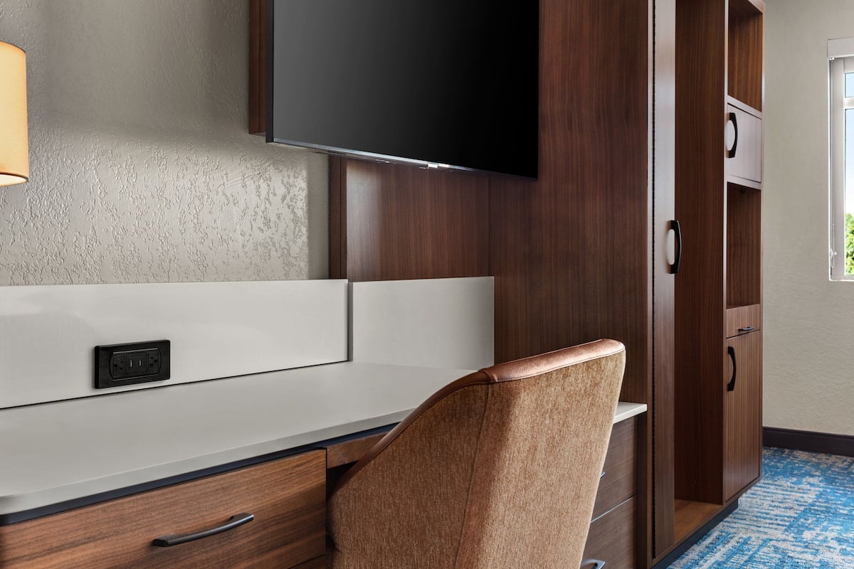 Habitación de hotel con televisión de pantalla plana montada en la pared, un escritorio con cajones, una silla y un armario con espejo.