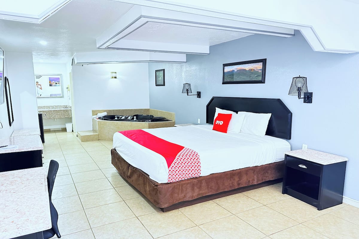 A hotel room with a bed, a small table, a chair, a nightstand, a wall-mounted lamp, and a picture on the wall.