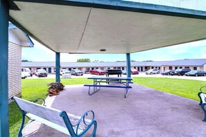 A covered outdoor area with picnic tables and benches, a brick building, and a parking lot with cars.