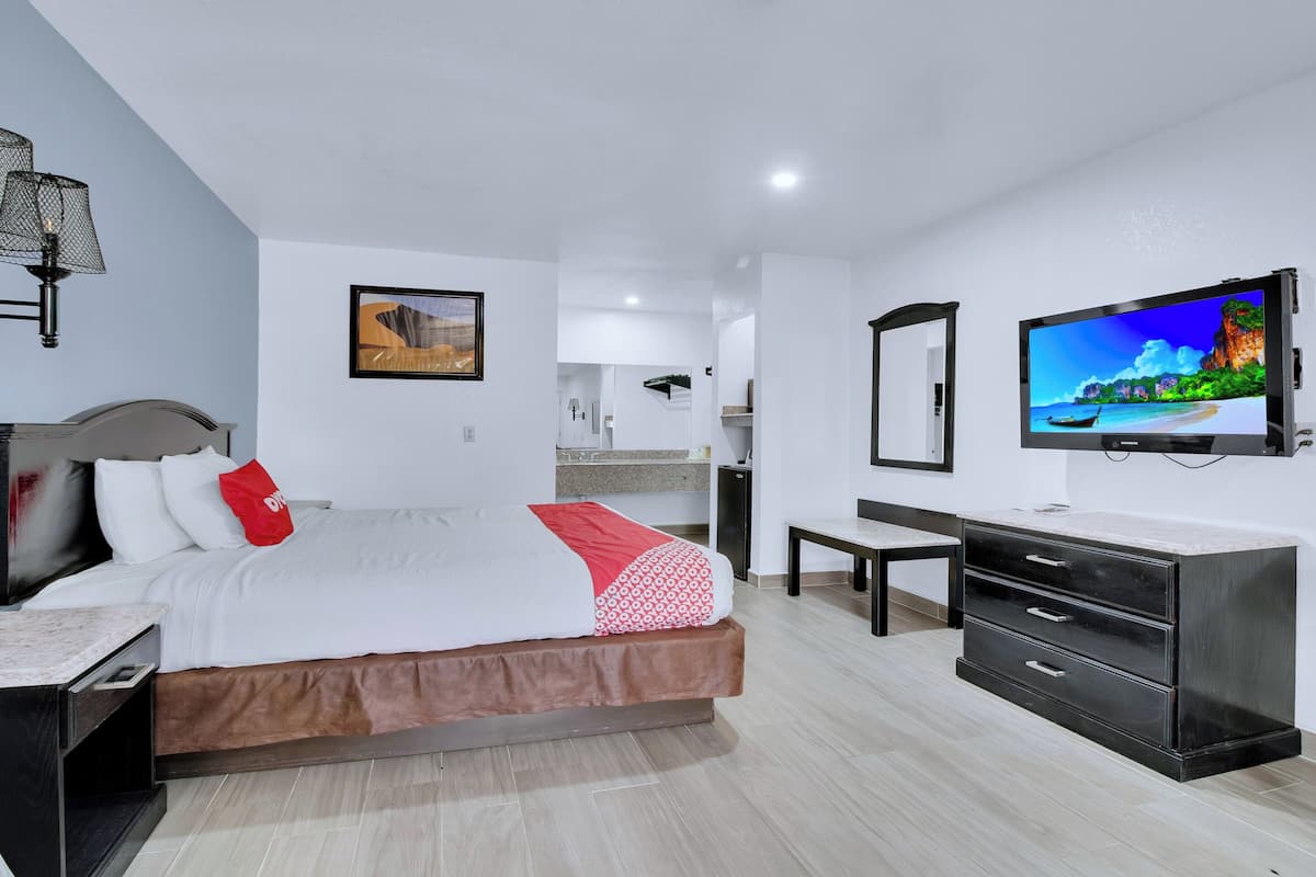 A modern hotel room with a bed, a desk, a dresser, a TV, and a bathroom visible in the background.