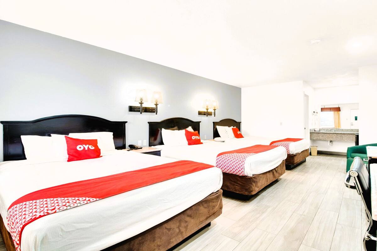A hotel room with three beds, each with a red and white bedspread and matching pillows. There are wall-mounted lights above each bed.