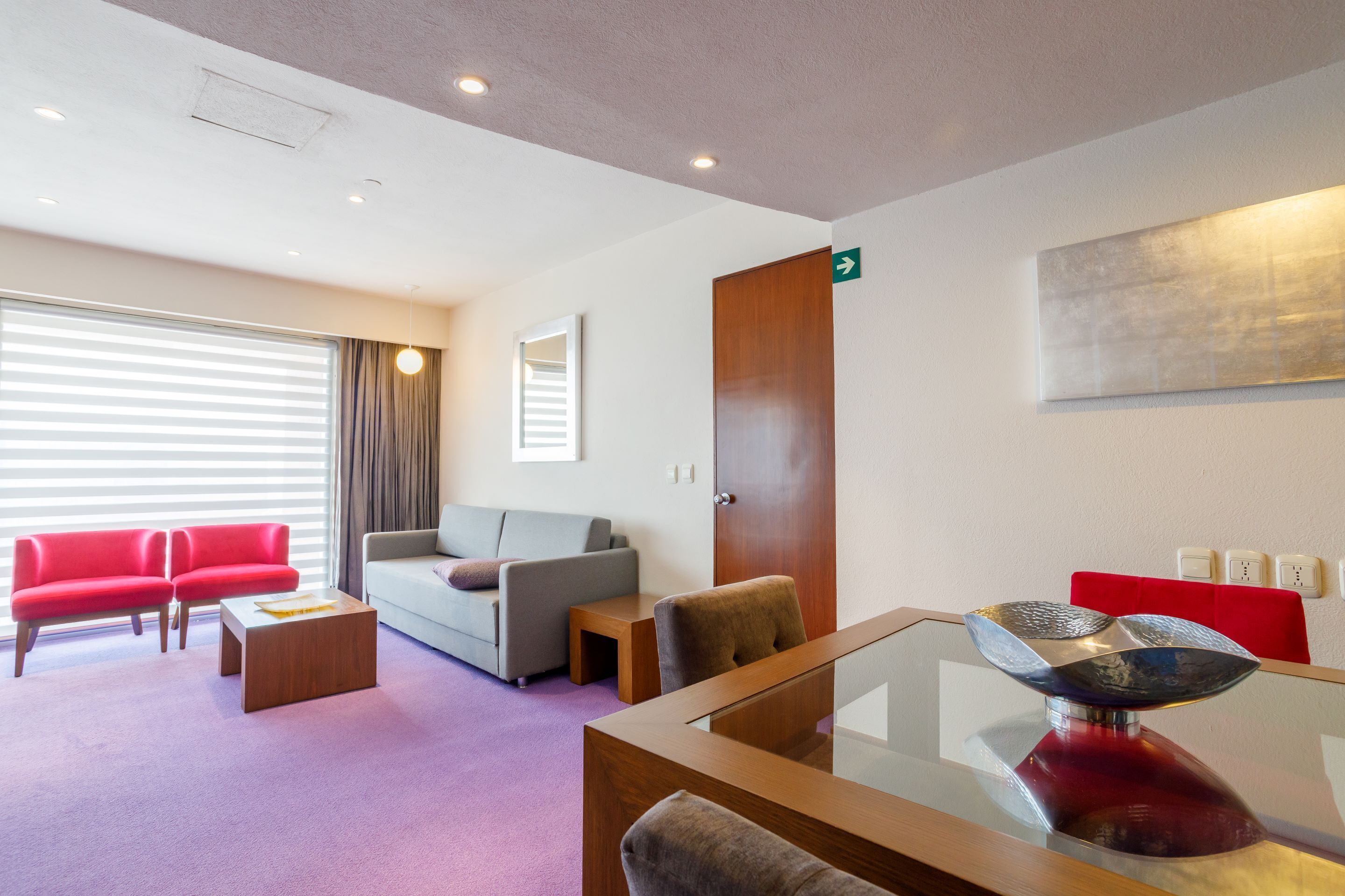 Un hall d’hôtel moderne avec un canapé rouge, un canapé gris et une table basse en verre sur laquelle est posé un bol décoratif.