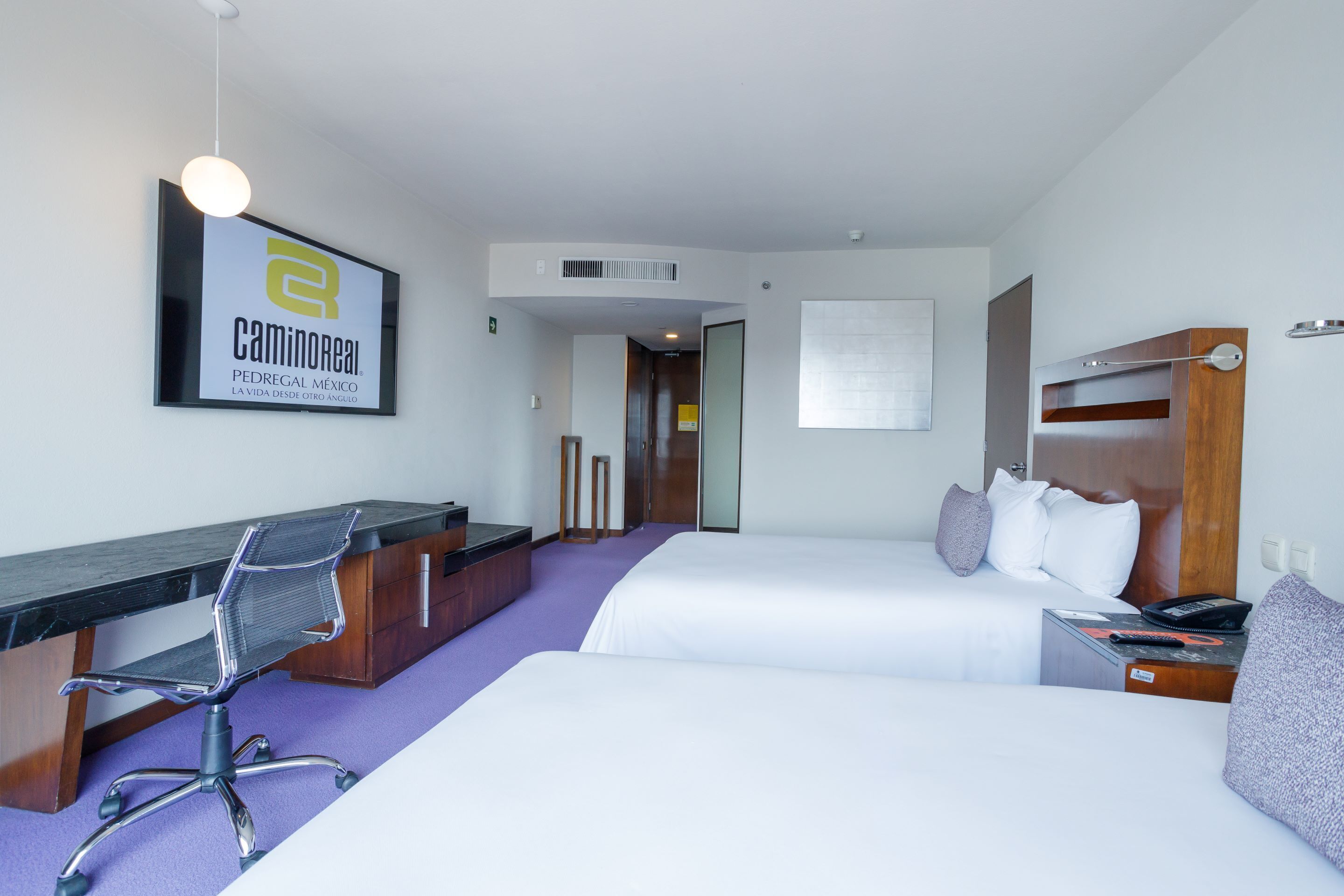 A hotel room with a desk, two beds, a chair, and a sign on the wall.