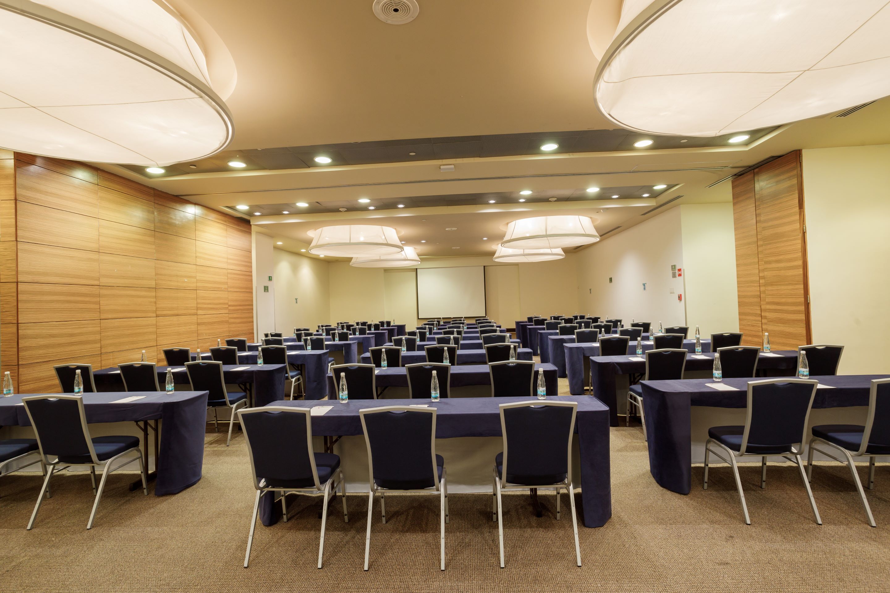A conference room with rows of tables and chairs, equipped with water bottles and a projector screen.