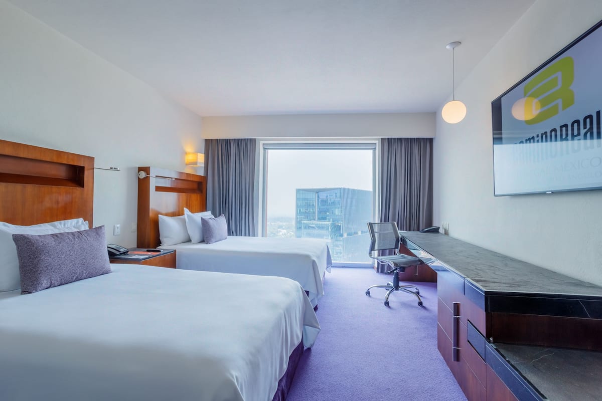 Ein Hotelzimmer mit zwei Betten, einem Schreibtisch und einem großen Fenster mit Blick auf die Stadt.