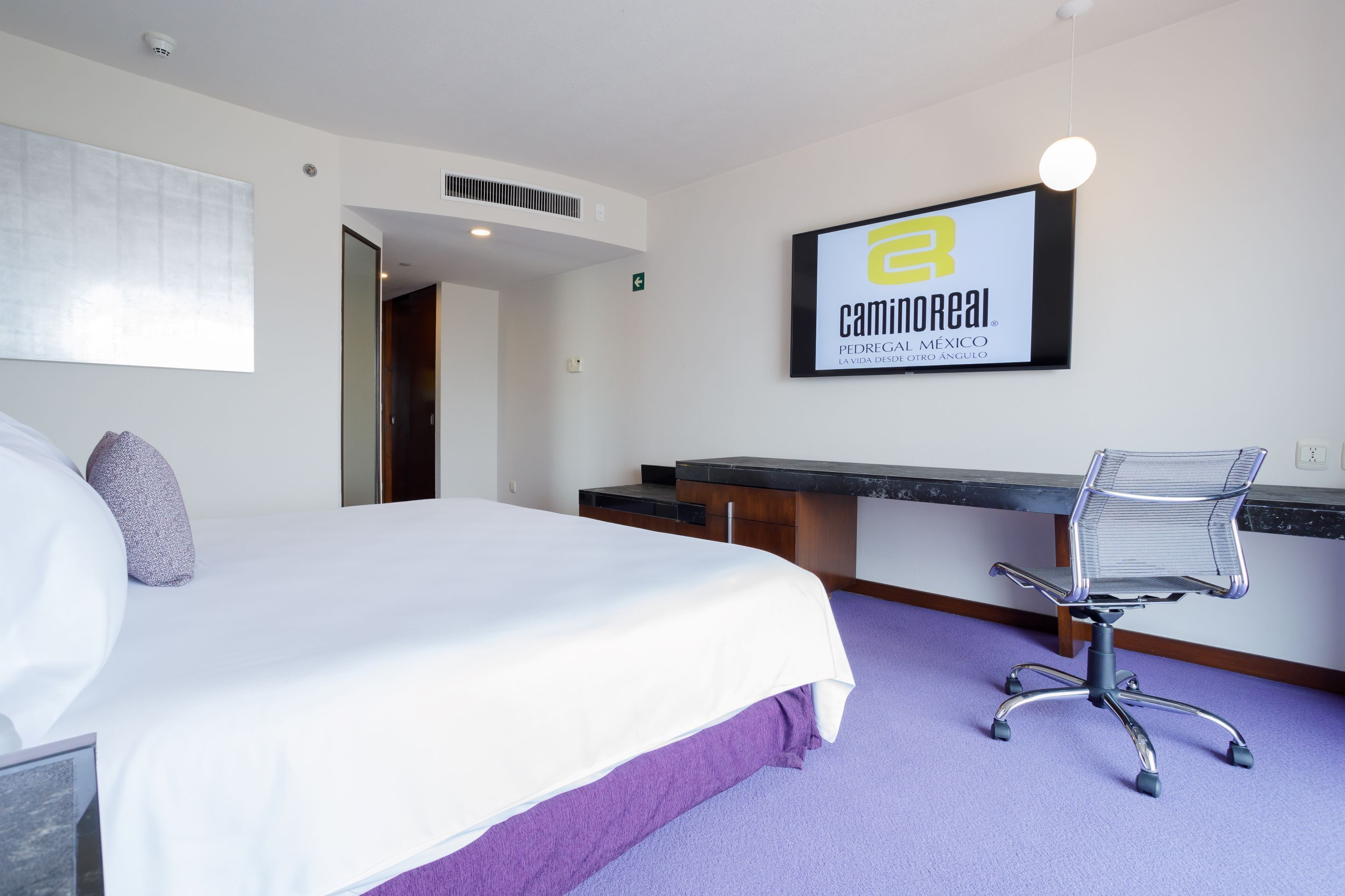 A hotel room with a bed, a desk, and a chair. There is a sign on the wall and a window with blinds.