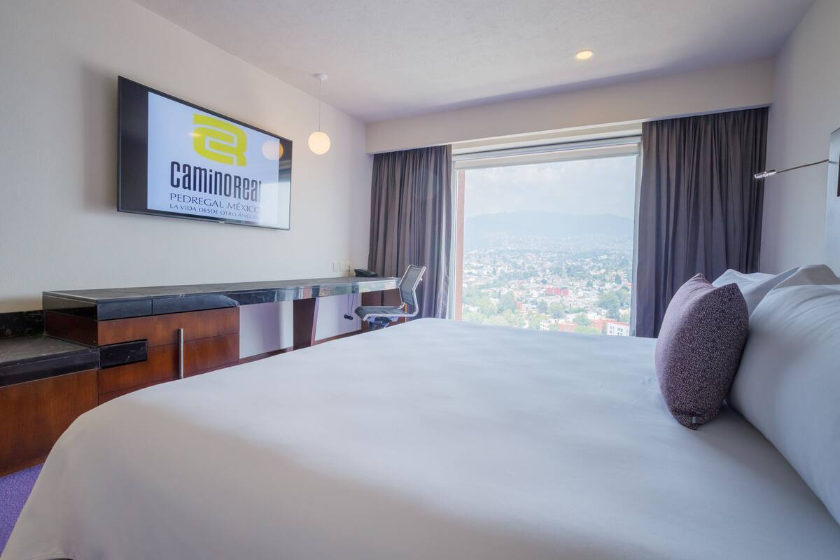 Ein Hotelzimmer mit einem großen Bett, einem Schreibtisch, einem Sessel und Blick auf die Stadt.