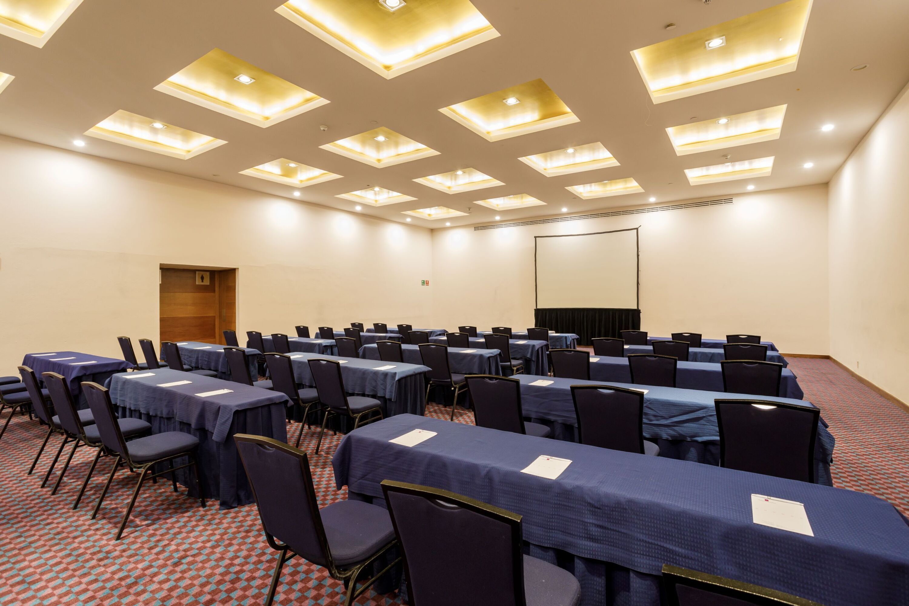 Un salón de conferencias con varias mesas y sillas, luces empotradas en el techo y una pantalla de proyección.
