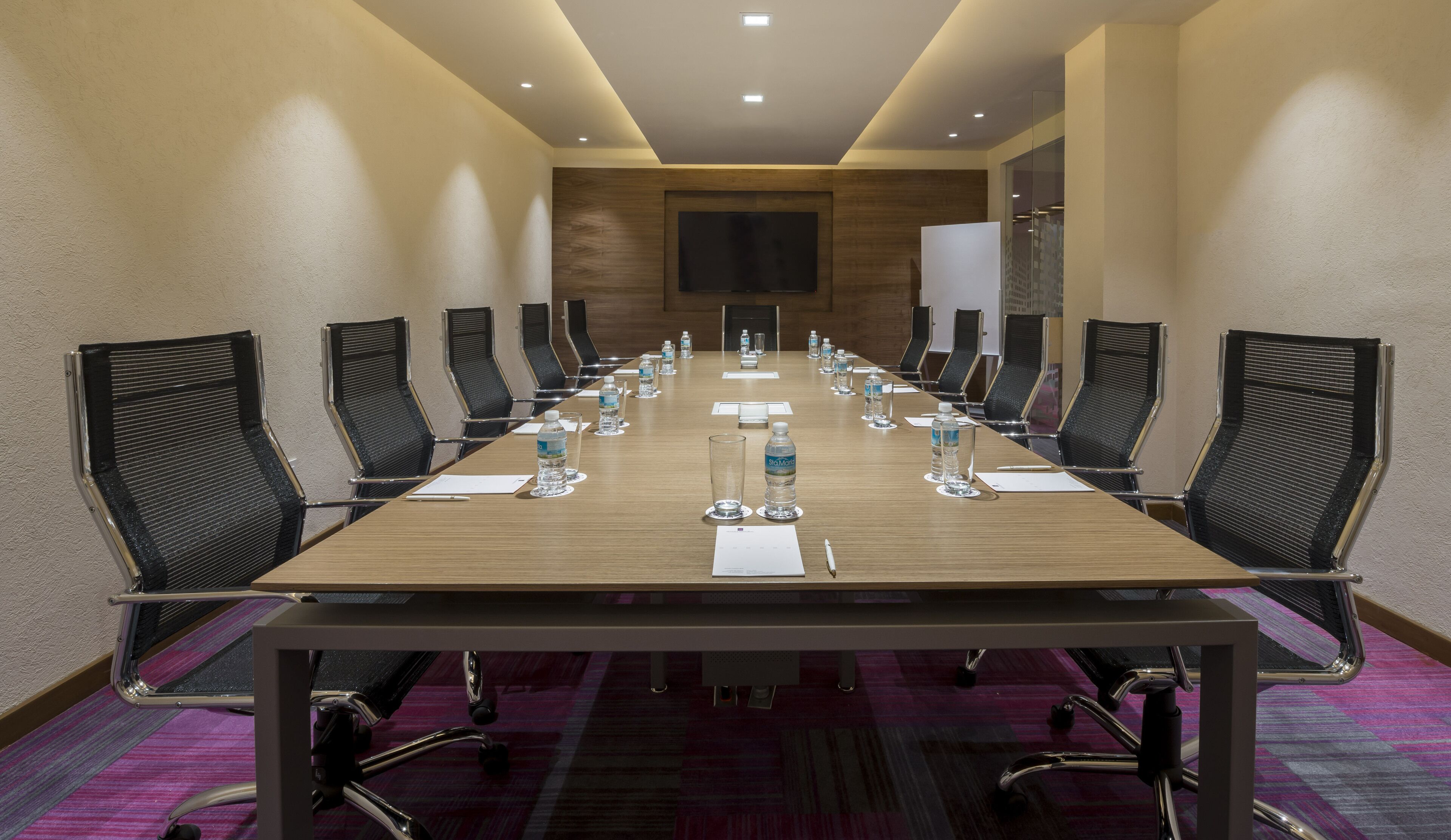 Une salle de conférence avec une longue table, des chaises noires, des bouteilles d’eau et des documents.