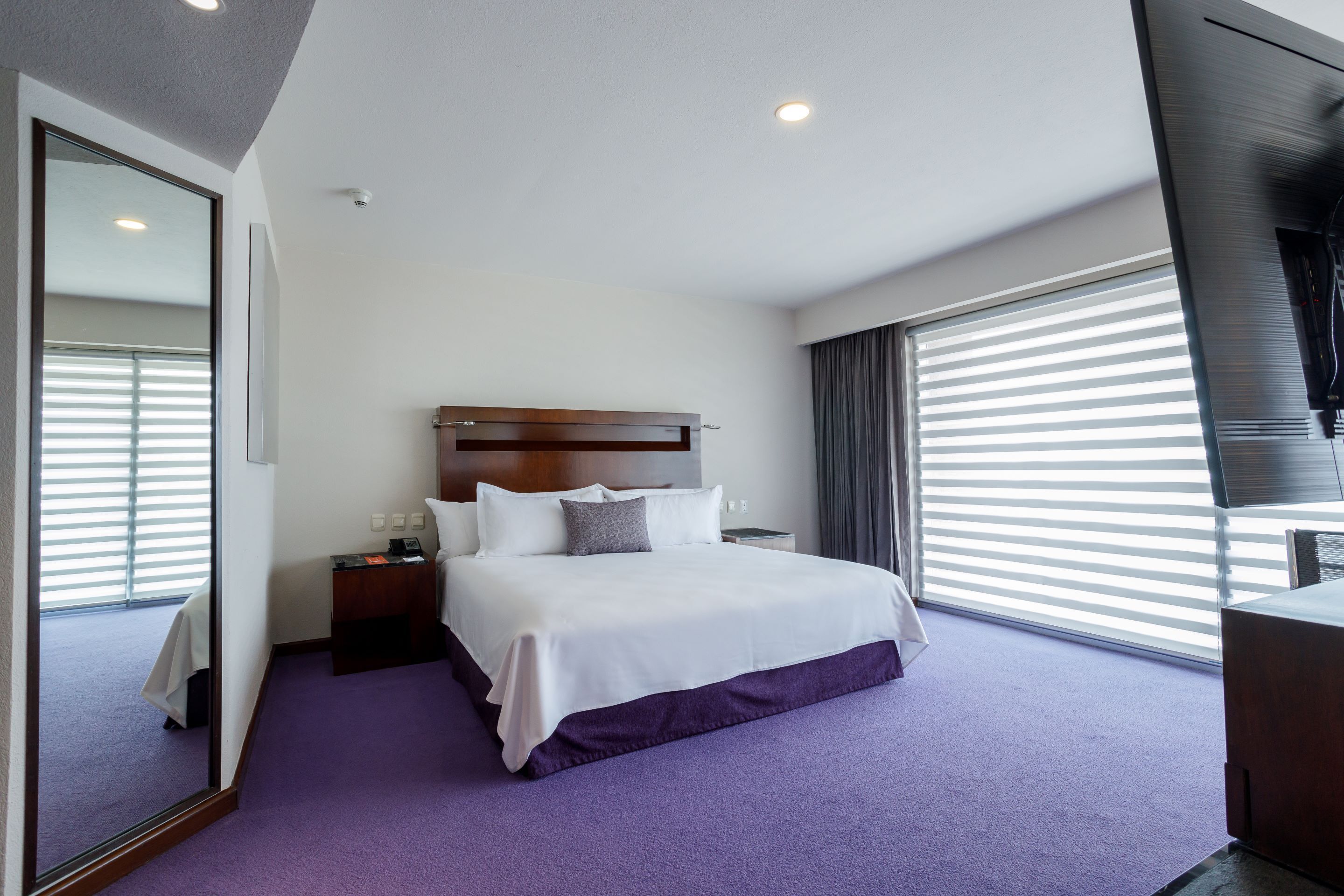 Ein Hotelzimmer mit einem großen Bett, einem Nachttisch, einem Fernseher und einem Fenster mit Jalousien.