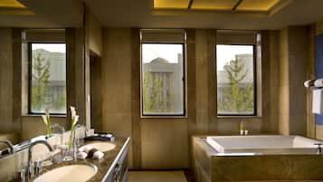 A modern bathroom with a large bathtub, a double sink vanity, and large windows offering views of buildings and trees.