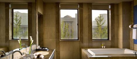 Un baño moderno con una bañera grande, un tocador doble y amplias ventanas que ofrecen vistas a edificios y árboles.