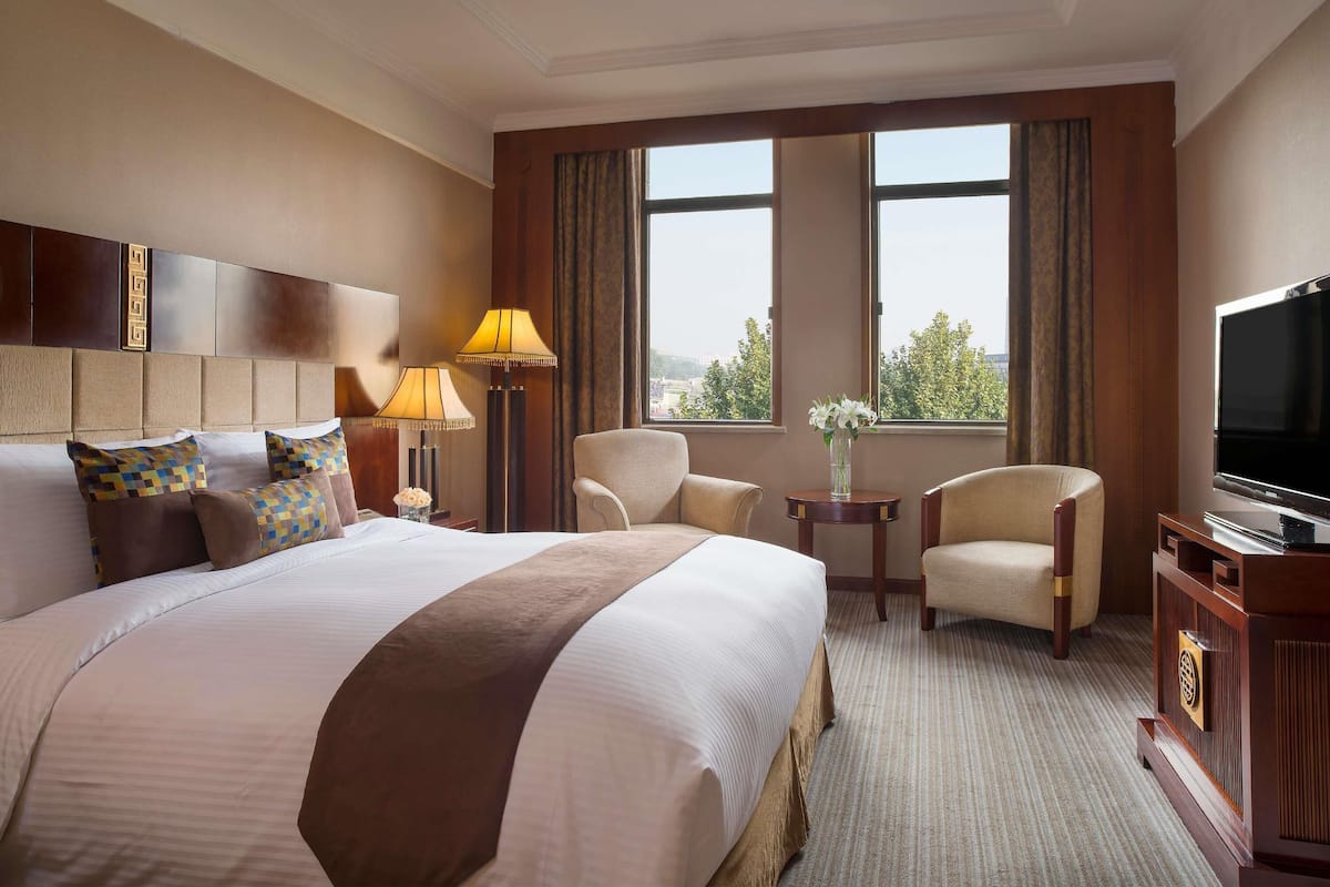 A hotel room with a large bed, two armchairs, a TV, and a window with a view.