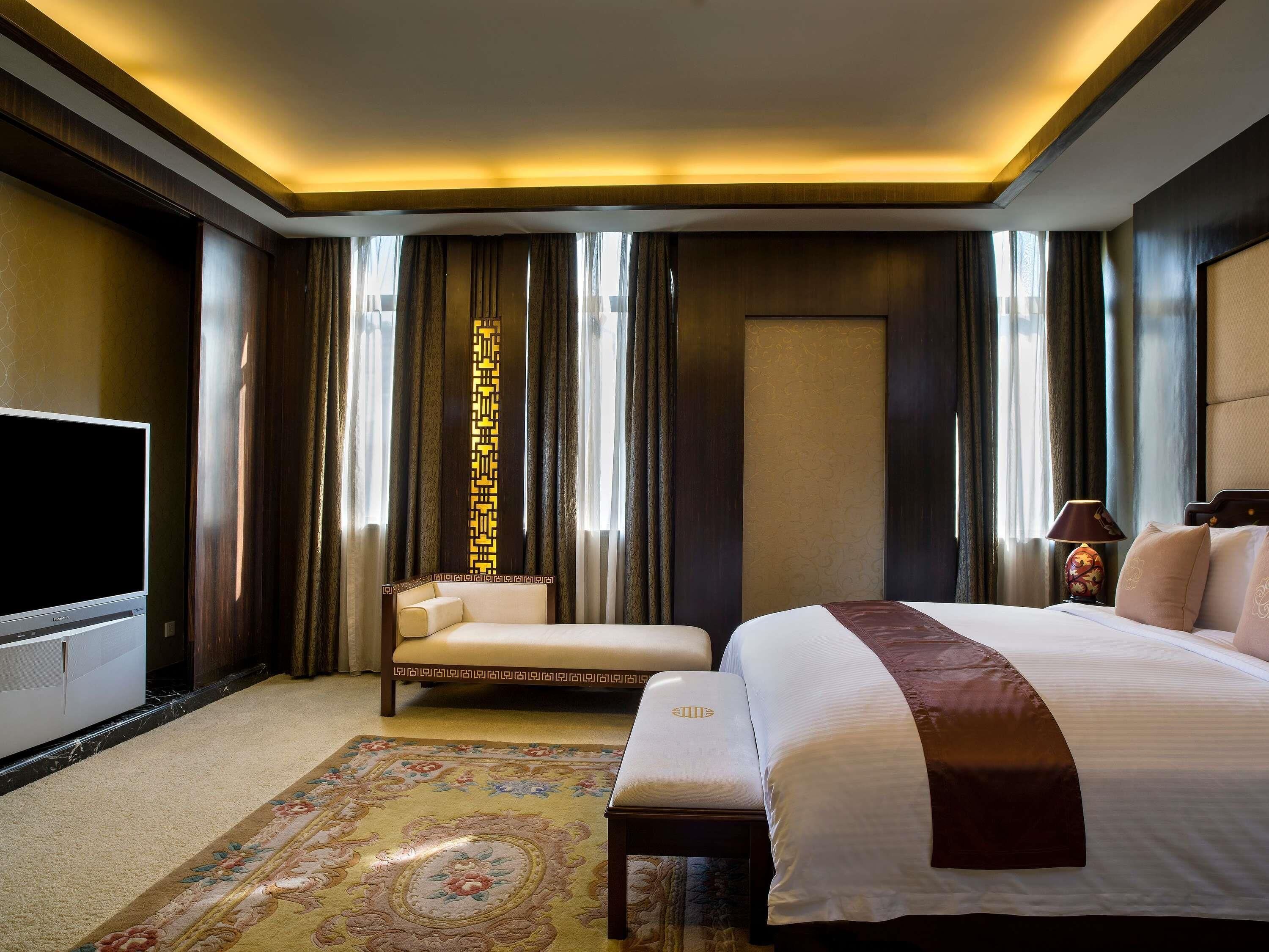 A hotel room with a large bed, a TV, a sofa, a decorative pillar, and a patterned rug.
