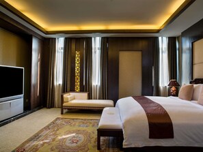 Ein Hotelzimmer mit einem großen Bett, einem Fernseher, einer Couch, einer Zierensäule und einem gemusterten Teppich.