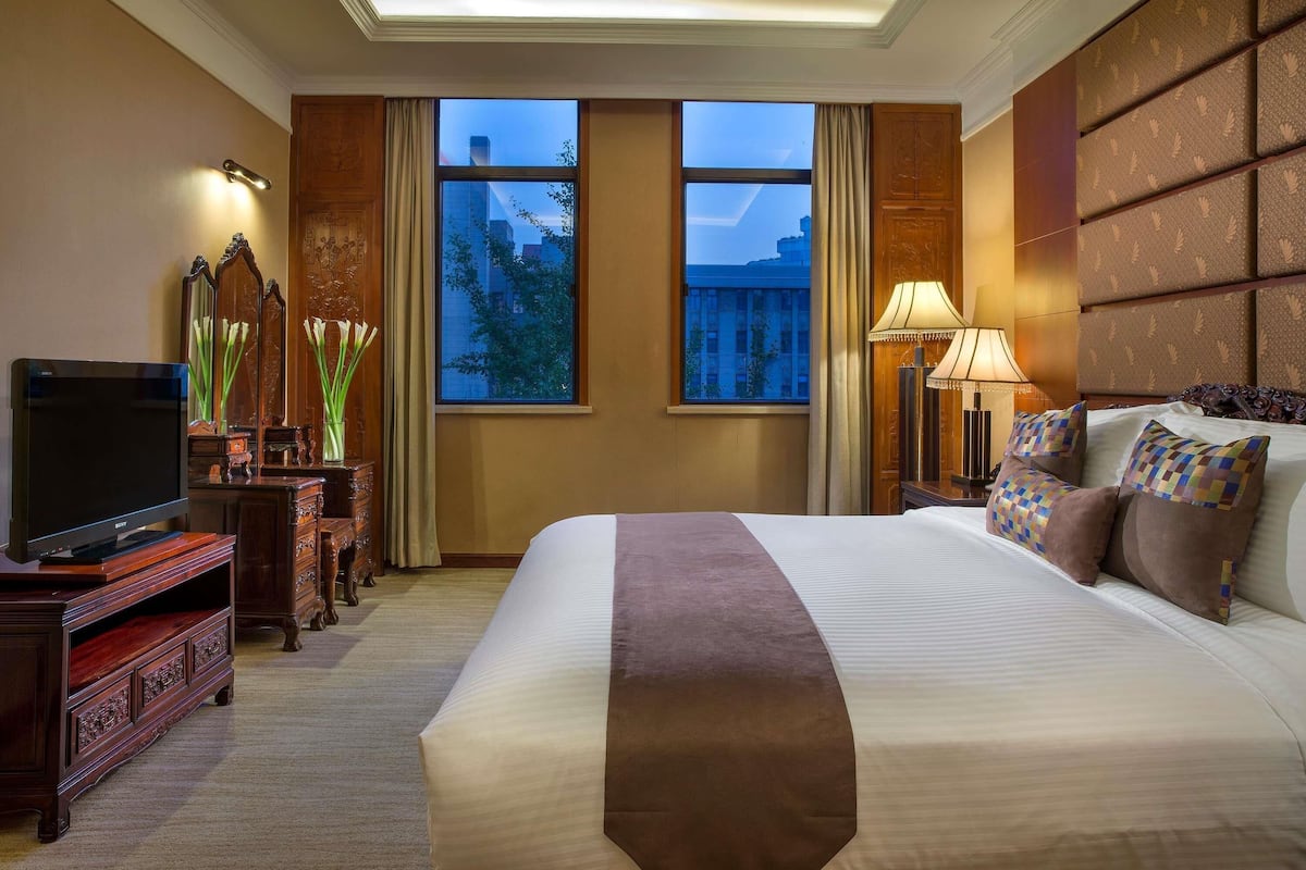 A hotel room with a large bed, a television, a desk with a lamp, and a view of the cityscape through the windows.