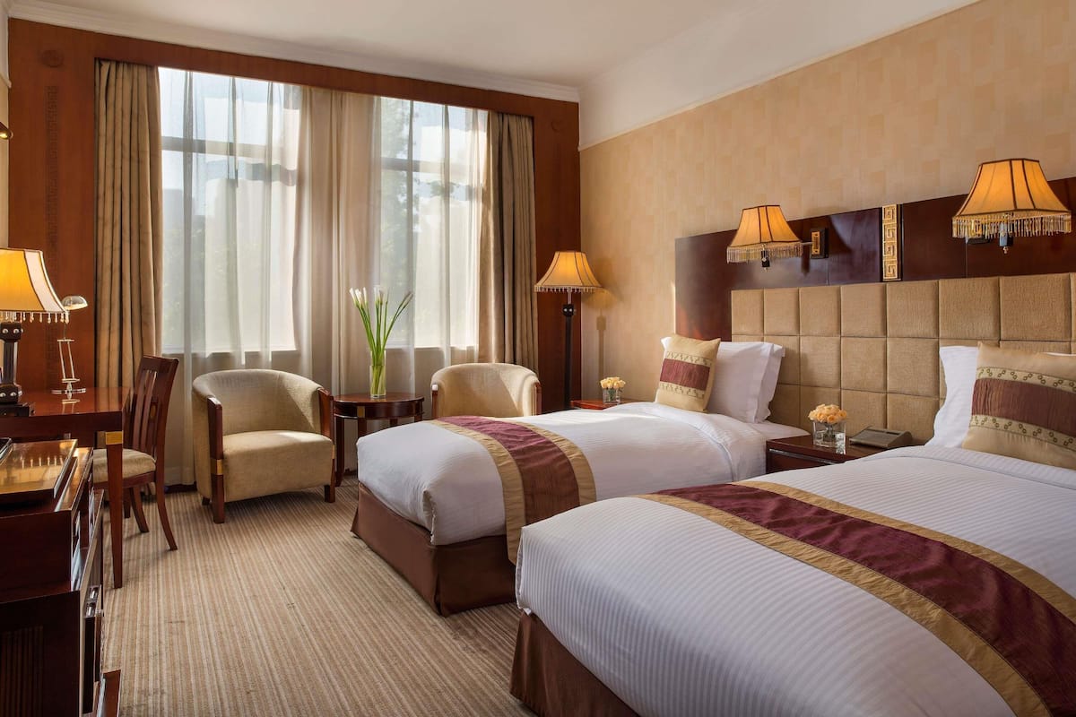 A hotel room with two beds, a desk with a lamp, a chair, and a large window with curtains.