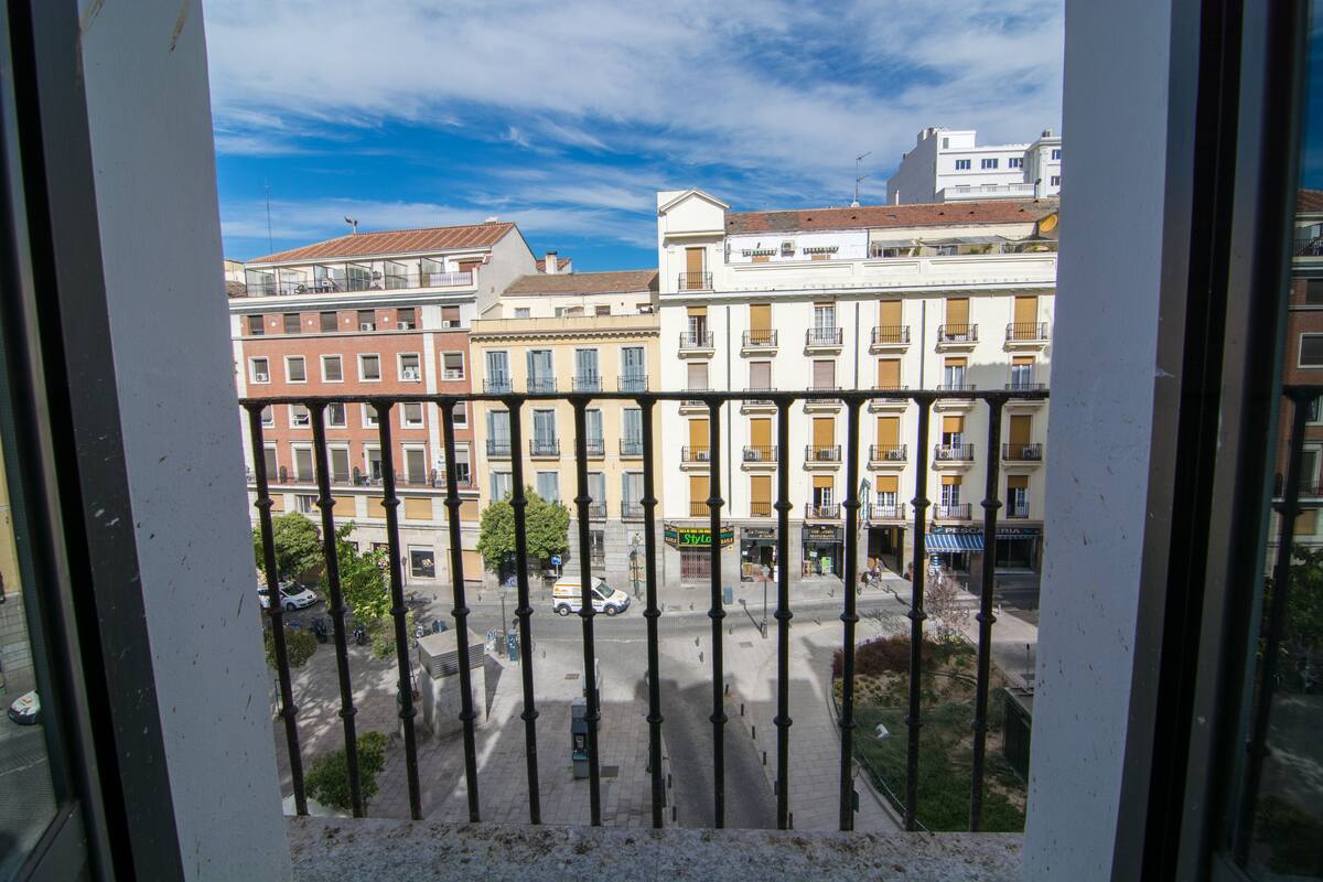 Vista a través de una ventana con baranda metálica, mostrando una calle con edificios, un árbol y un coche estacionado.